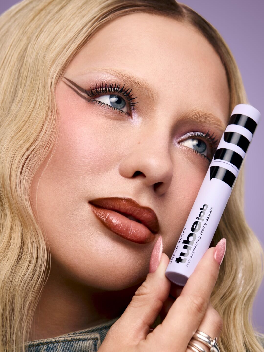 Close up of a model with graphic liner holding up a mascara to their cheek against a lavender backdrop
